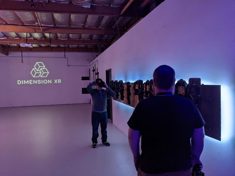 A player in a headset under the glowing Dimension XR logo in the first warehouse arena, beside a lit rack of blasters.