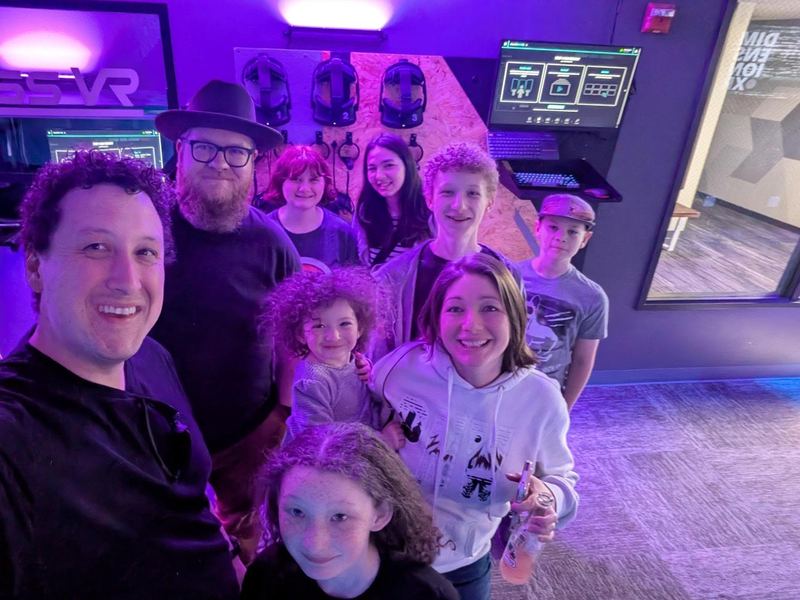 A group selfie of friends and family in the purple-lit arena, with headsets on the wall behind them.