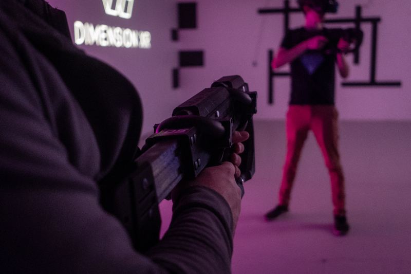 Seen over a player’s shoulder, an opponent aims a blaster in front of the Dimension XR logo in the purple-lit Tukwila arena.