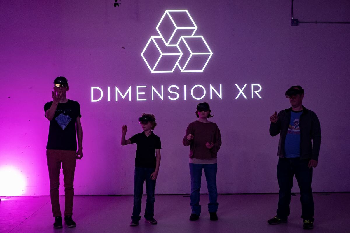 Four players with headsets pushed up on their foreheads stand in front of the glowing Dimension XR logo on the arena wall in Tukwila, 2022.
