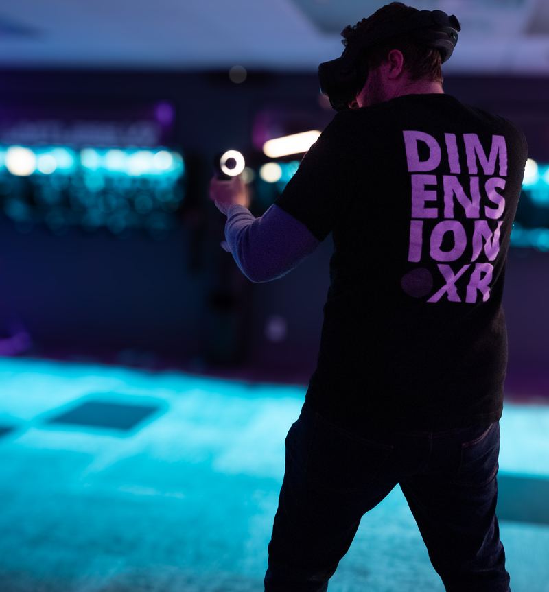 A player in a Dimension XR T-shirt aims a controller across the blue-lit arena.